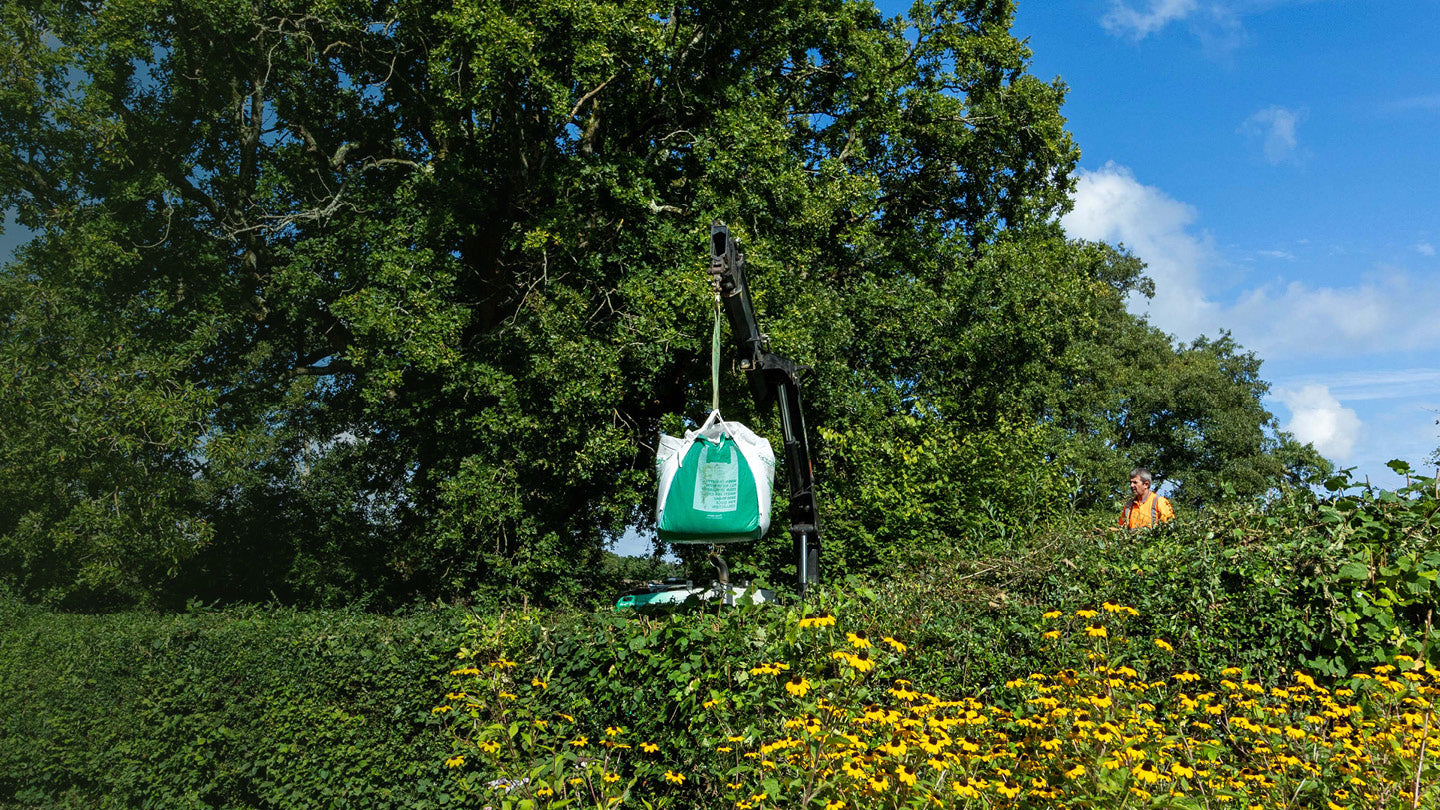 A green and white bulk bag suspended from a crane stand in a garden setting with trees, blue sky, and yellow flowers.